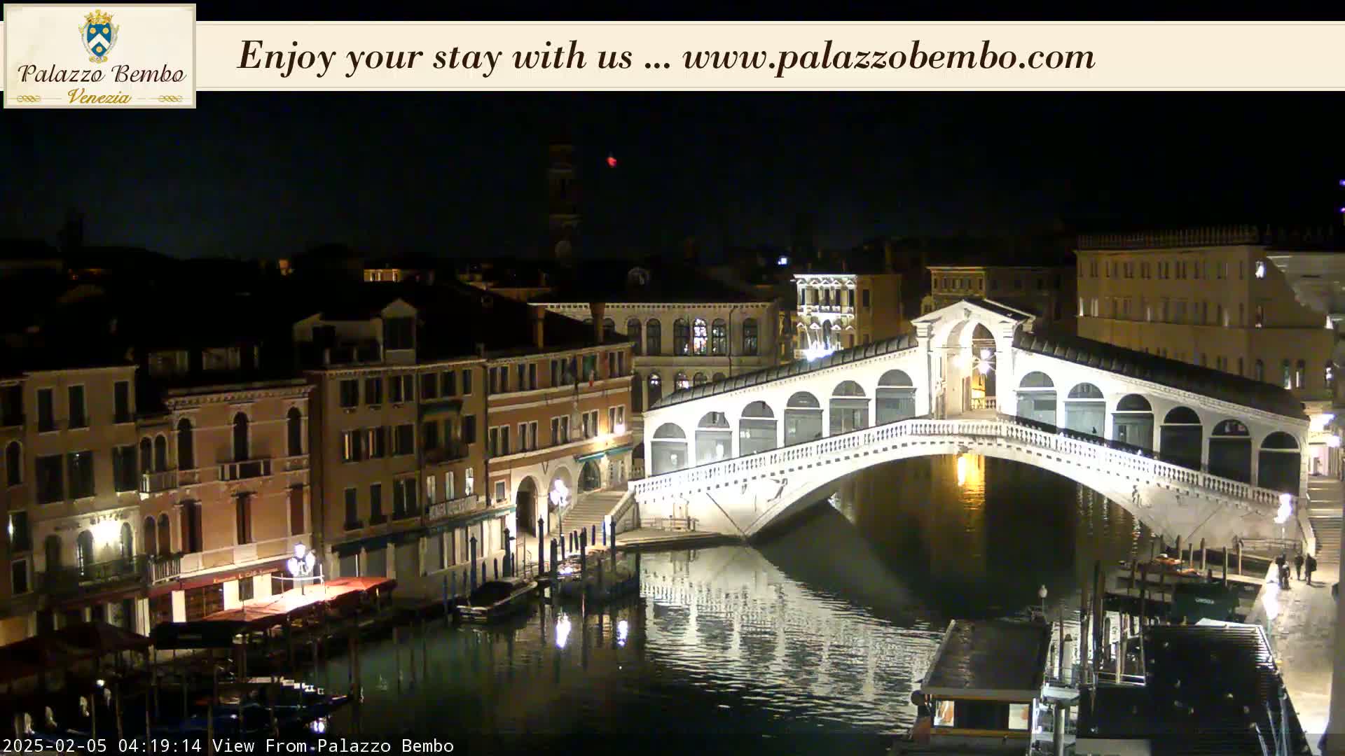 A nighttime view of a canal in Venice, showing the Rialto Bridge lit up, with buildings lining the waterway.