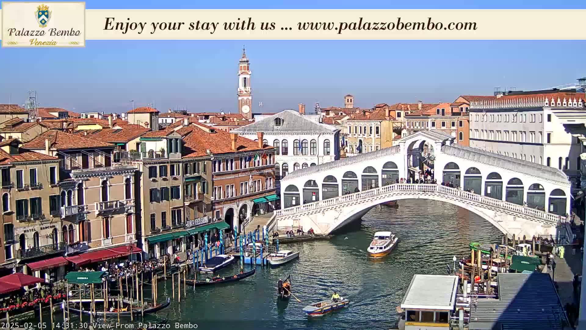 A sunny day in Venice shows a canal with boats, a white bridge with people on it, and numerous buildings lining the waterway.