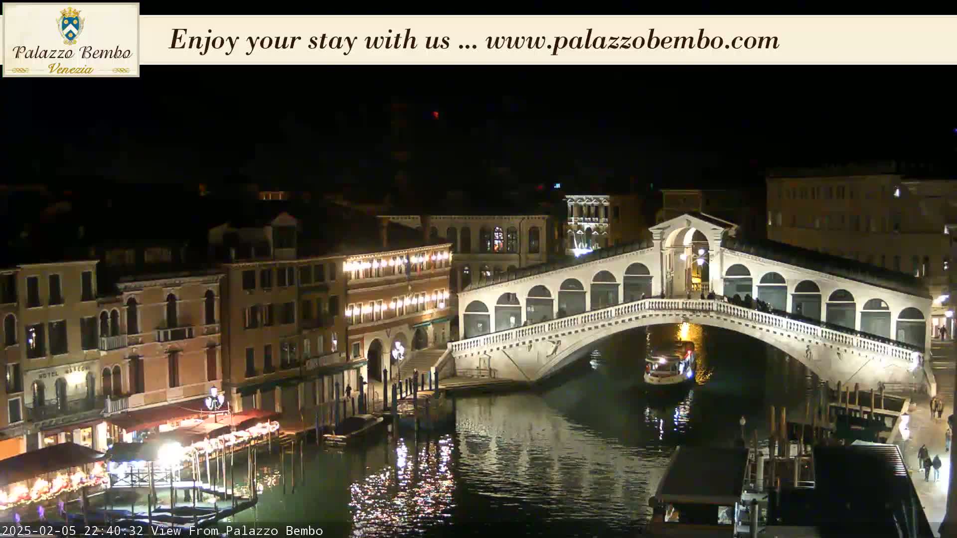 A nighttime view of a canal in Venice, showing a bridge illuminated with lights and buildings along the waterway.