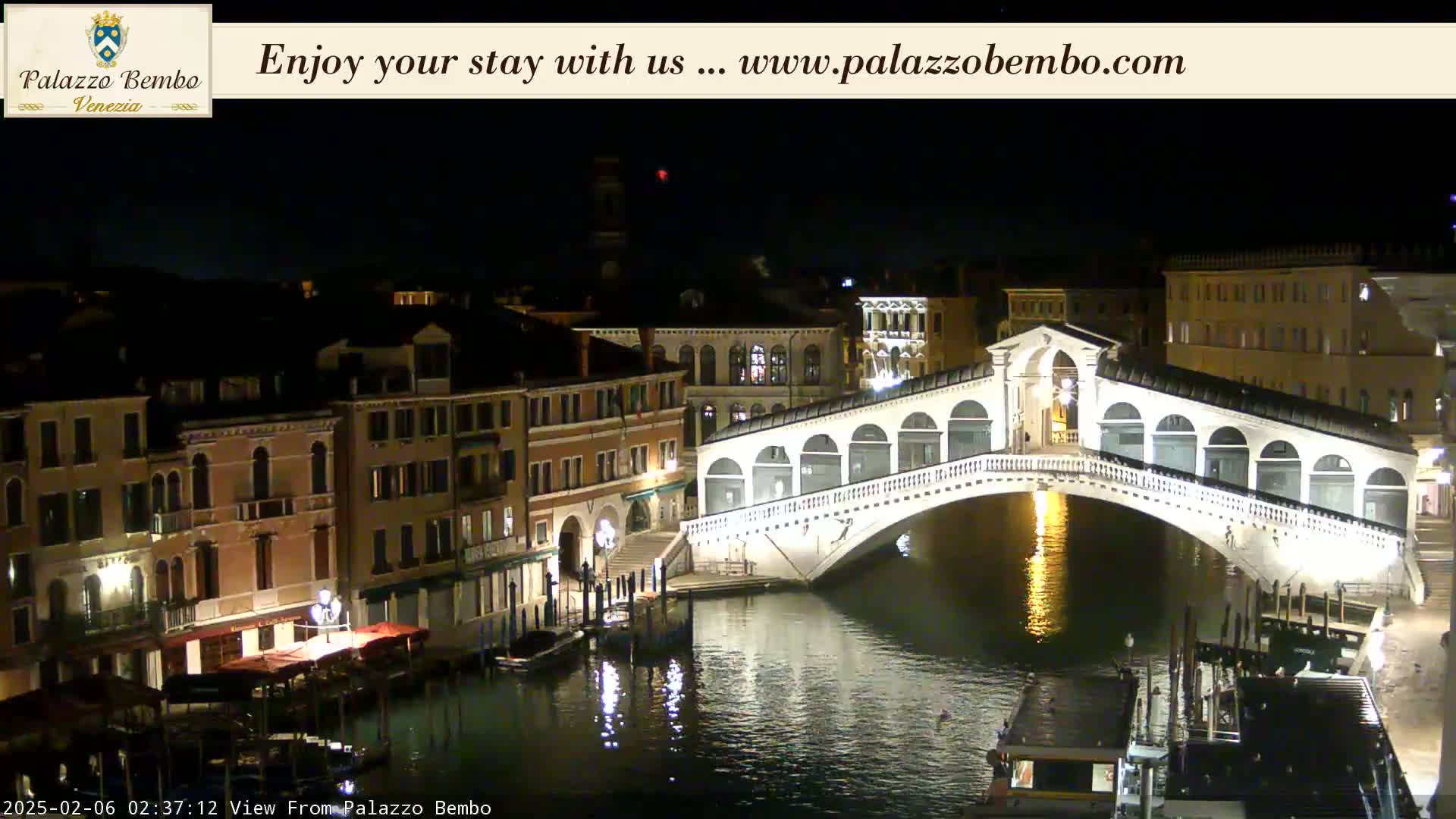 A nighttime view of a canal in Venice, with the Rialto Bridge illuminated and boats moored along the waterway, under a clear sky.