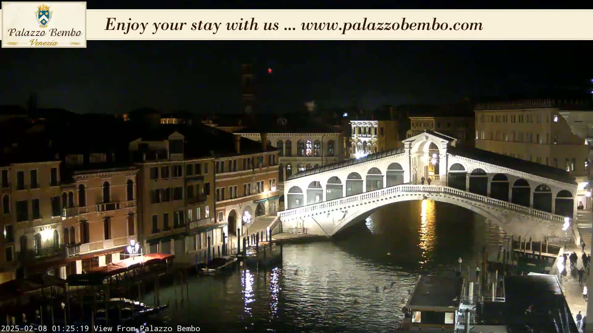 A nighttime view of a canal in Venice, showing a bridge lit up against the dark sky and buildings lining the waterway.