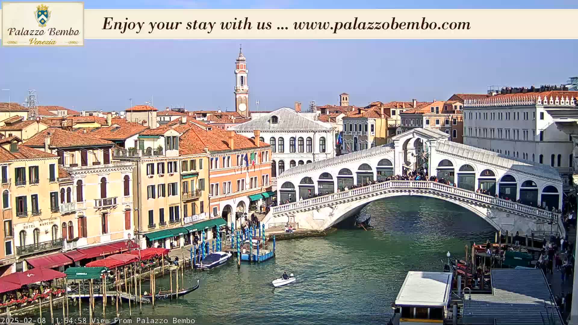 A sunny Venetian cityscape shows a canal with gondolas and a bridge crowded with people, with colorful buildings lining the waterway.