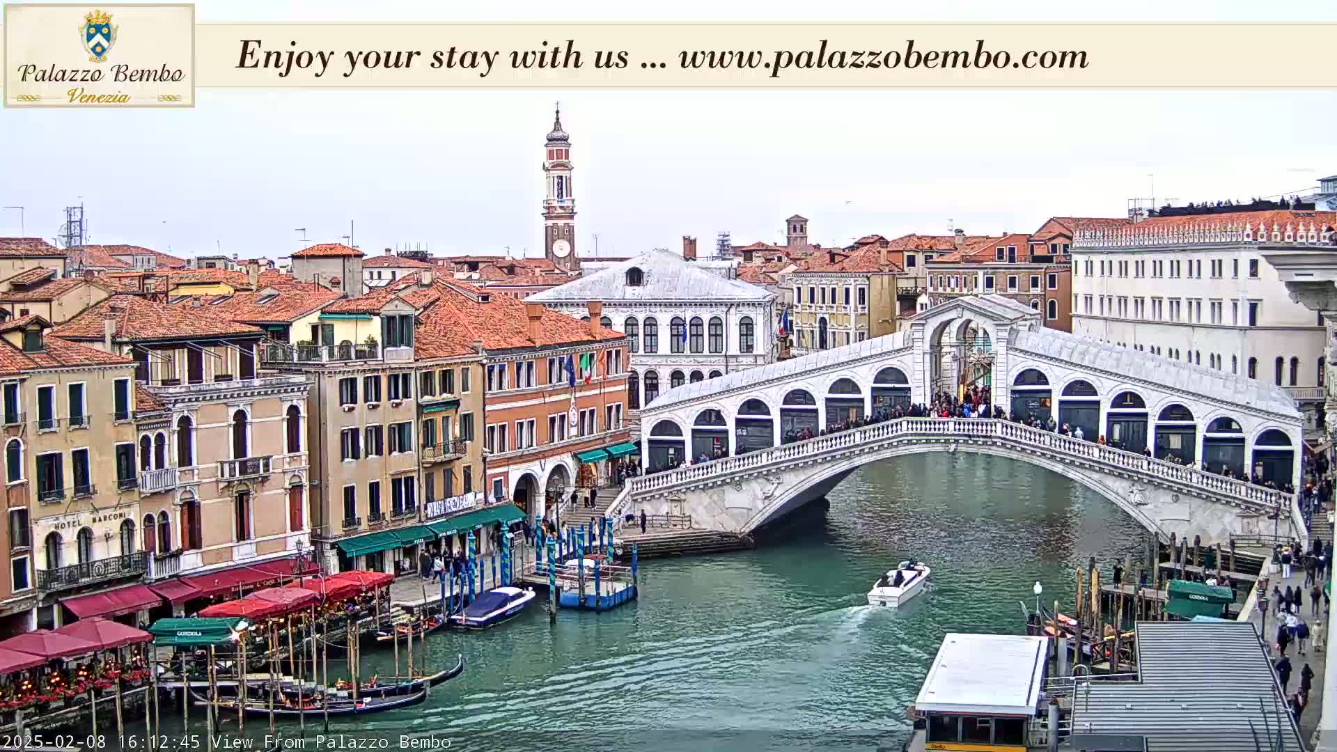 A view of a canal in Venice, under a cloudy sky, shows a bridge filled with people, gondolas, and boats on the water, and numerous buildings lining the canal.