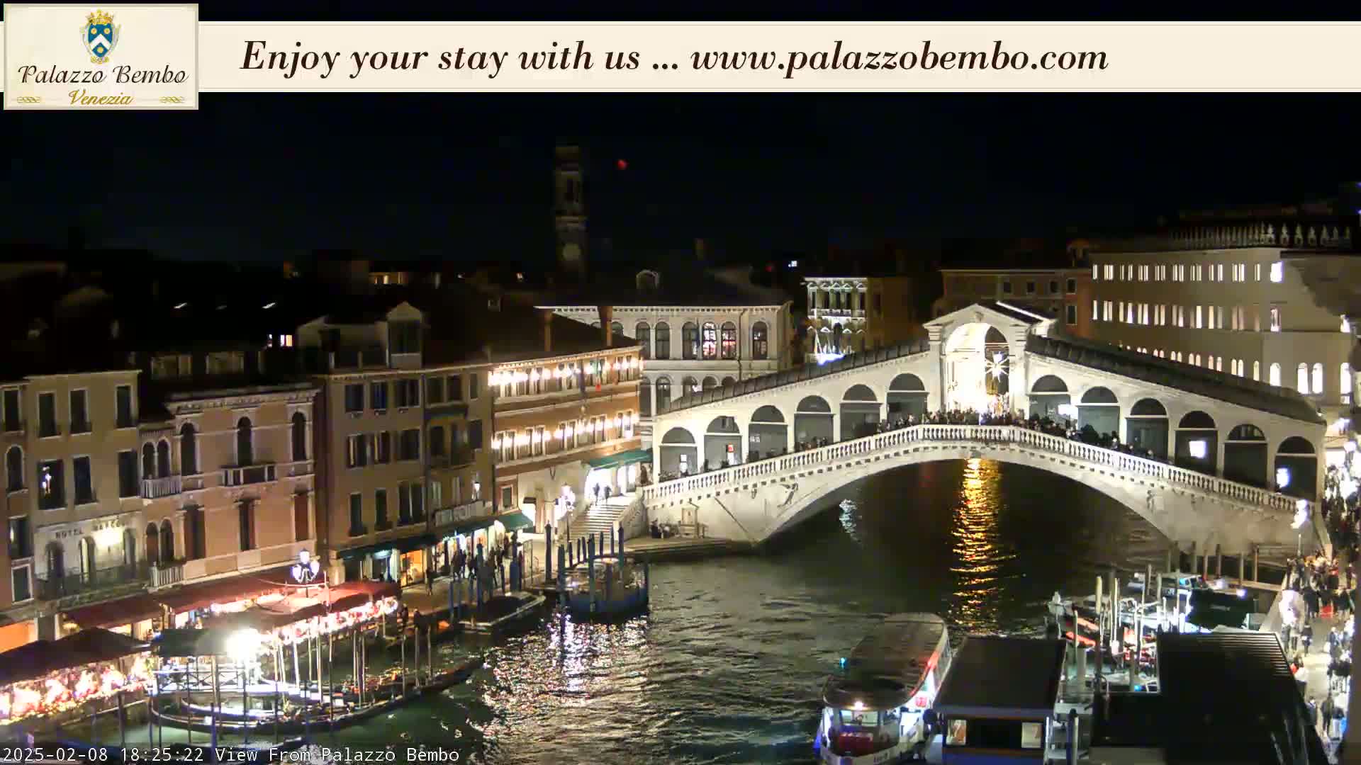 A nighttime view of a canal in Venice, showing the Rialto Bridge illuminated and crowded with people, with boats moored along the canal's edge.