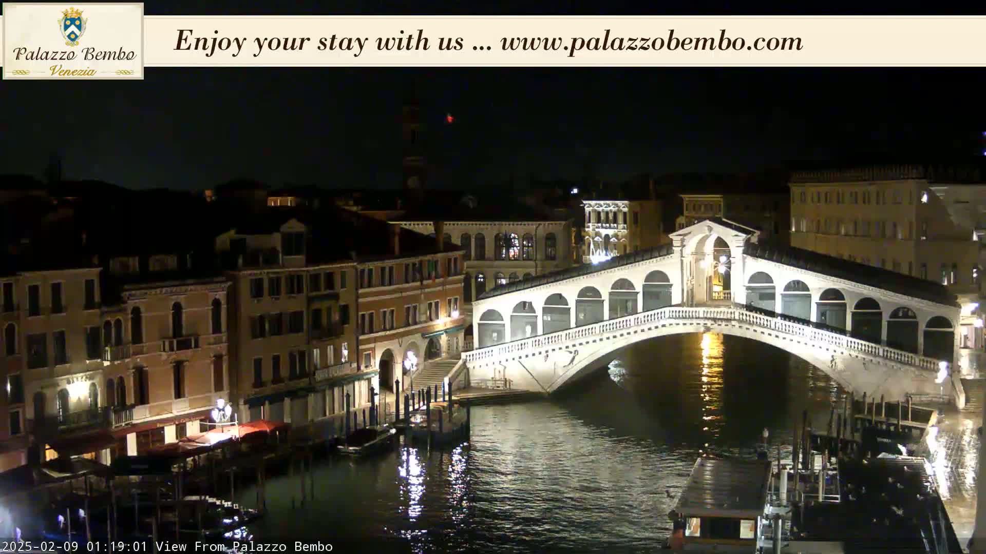 A nighttime view of a canal in Venice, showing a bridge lit up and buildings along the water's edge.
