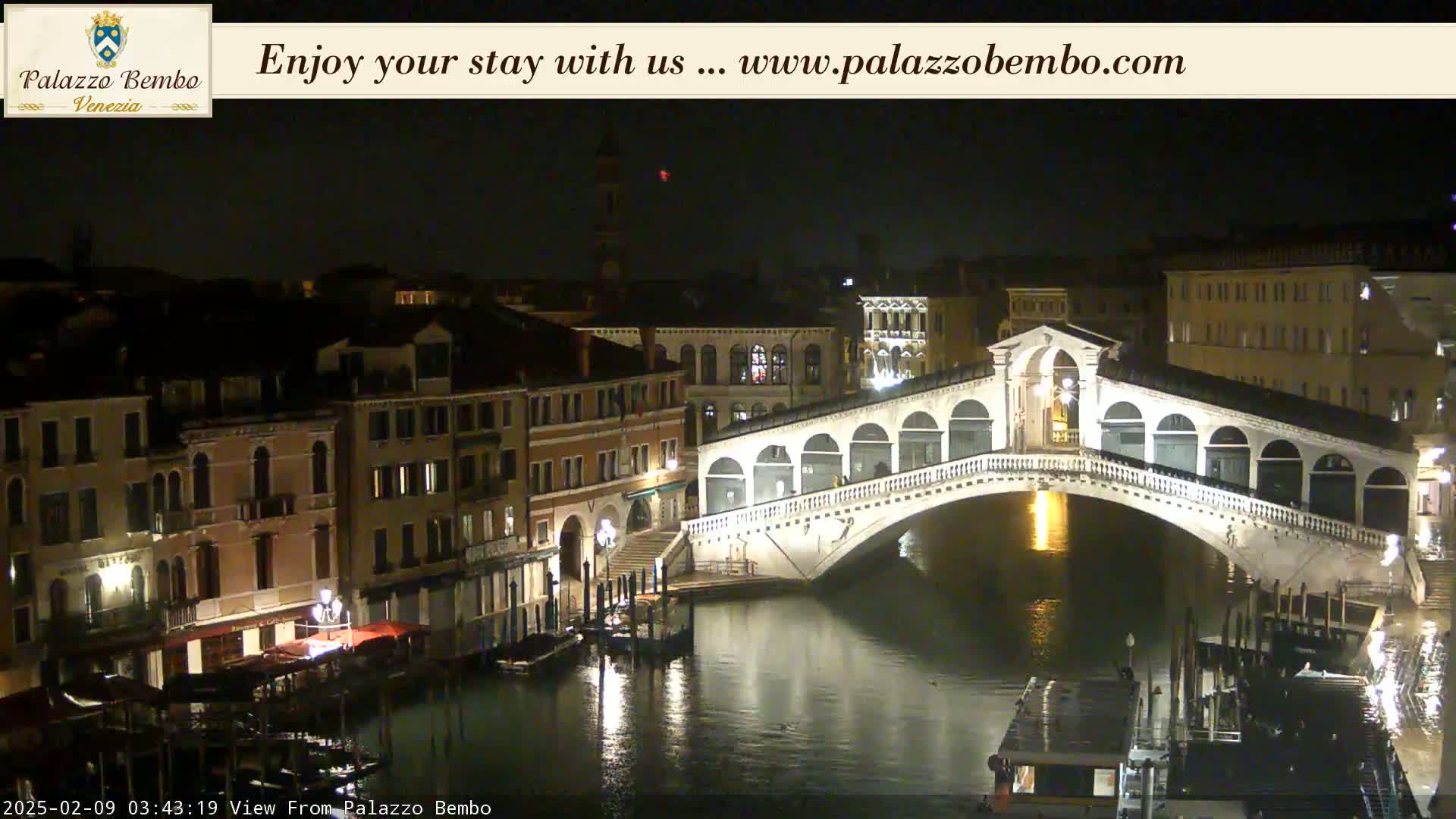 A nighttime view of a canal in Venice, showing a bridge illuminated, with buildings lining the waterway.
