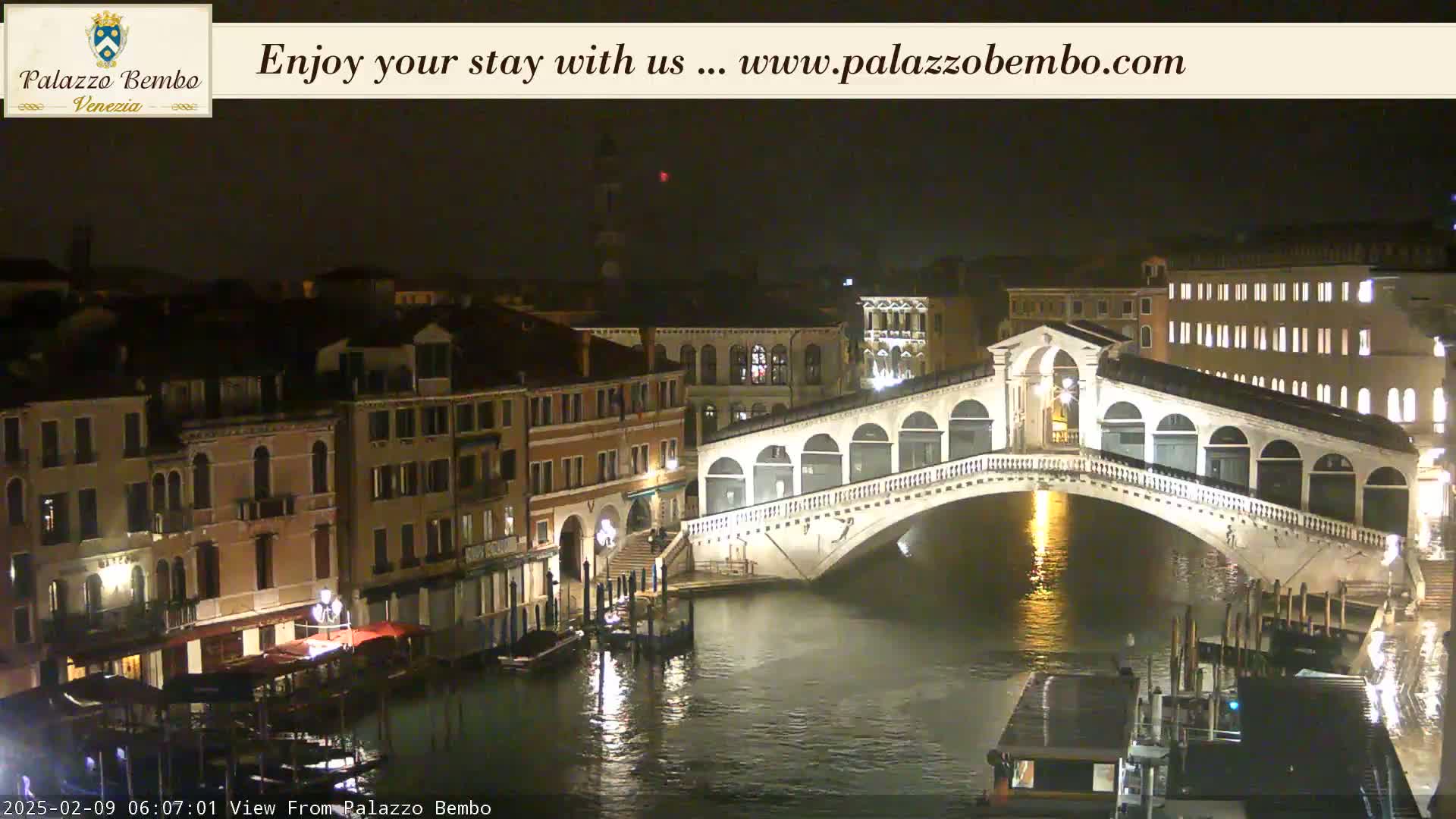 A nighttime view of a bridge spanning a canal in Venice, with buildings illuminated along the waterway.
