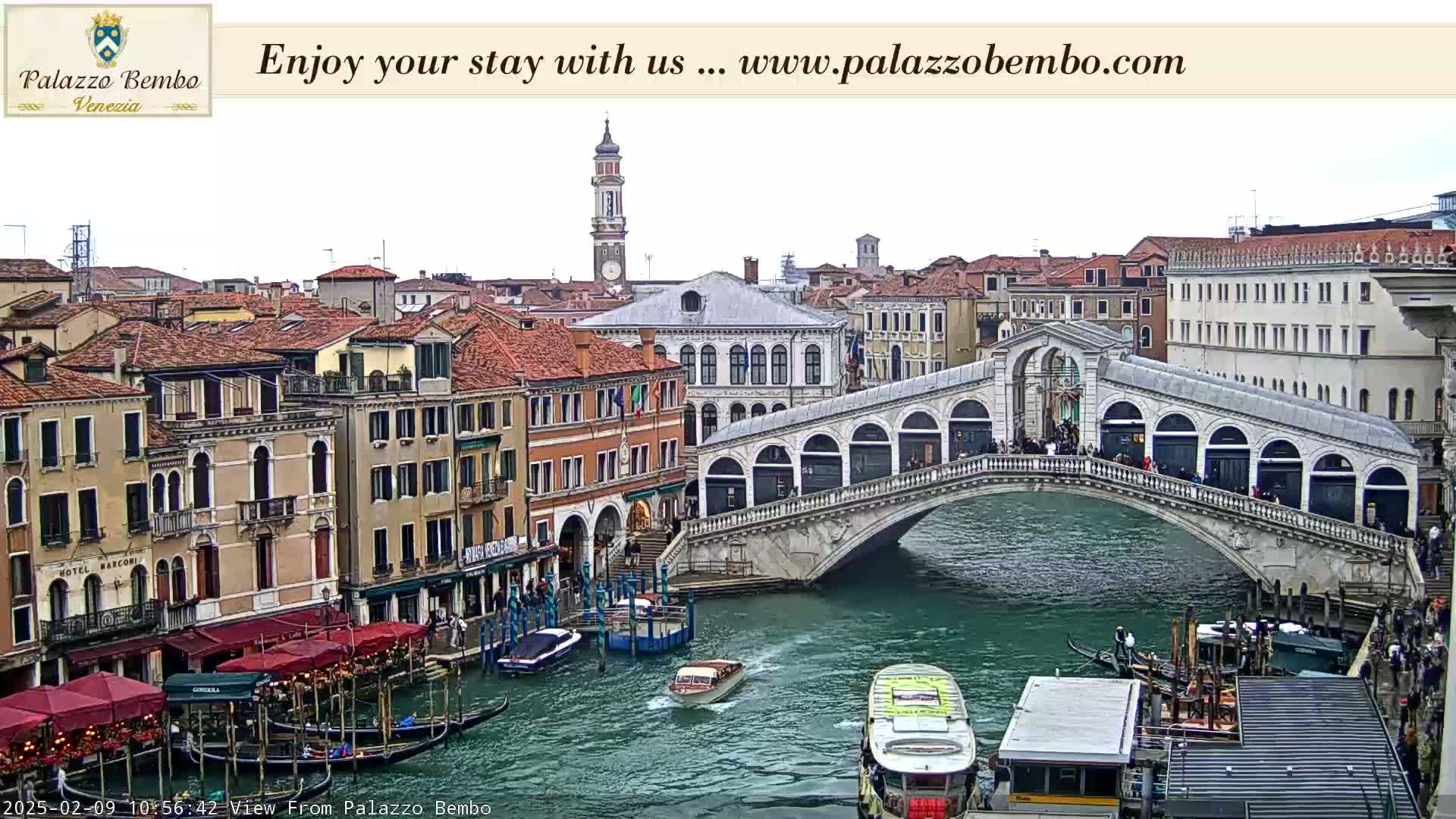 A canal in Venice, under a cloudy sky, shows a bridge with people walking across it, gondolas in the water, and buildings lining the waterway.