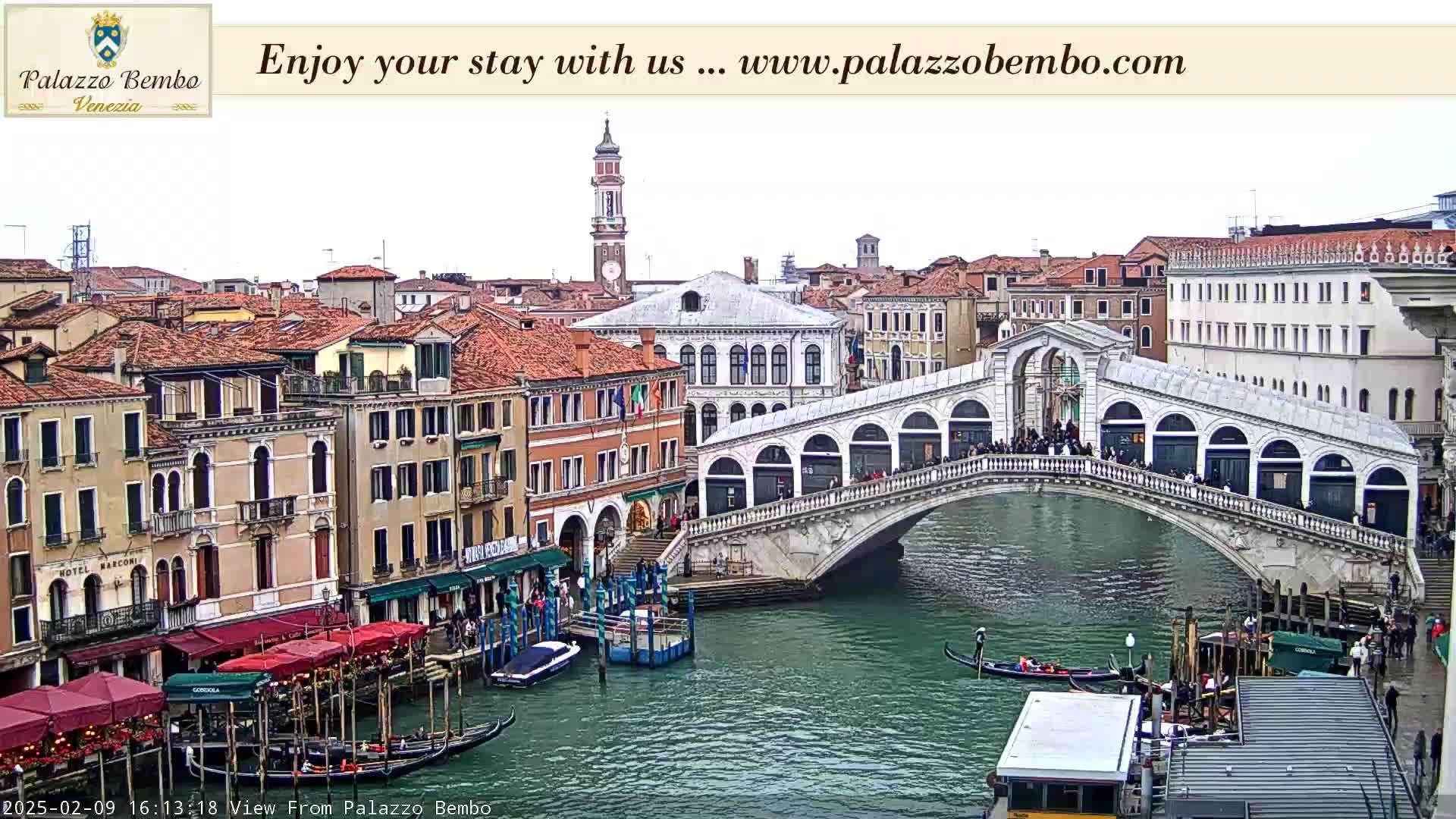 A view of a canal in Venice, on an overcast day, shows the Rialto Bridge with people walking across it, gondolas in the water, and colorful buildings lining the canal.
