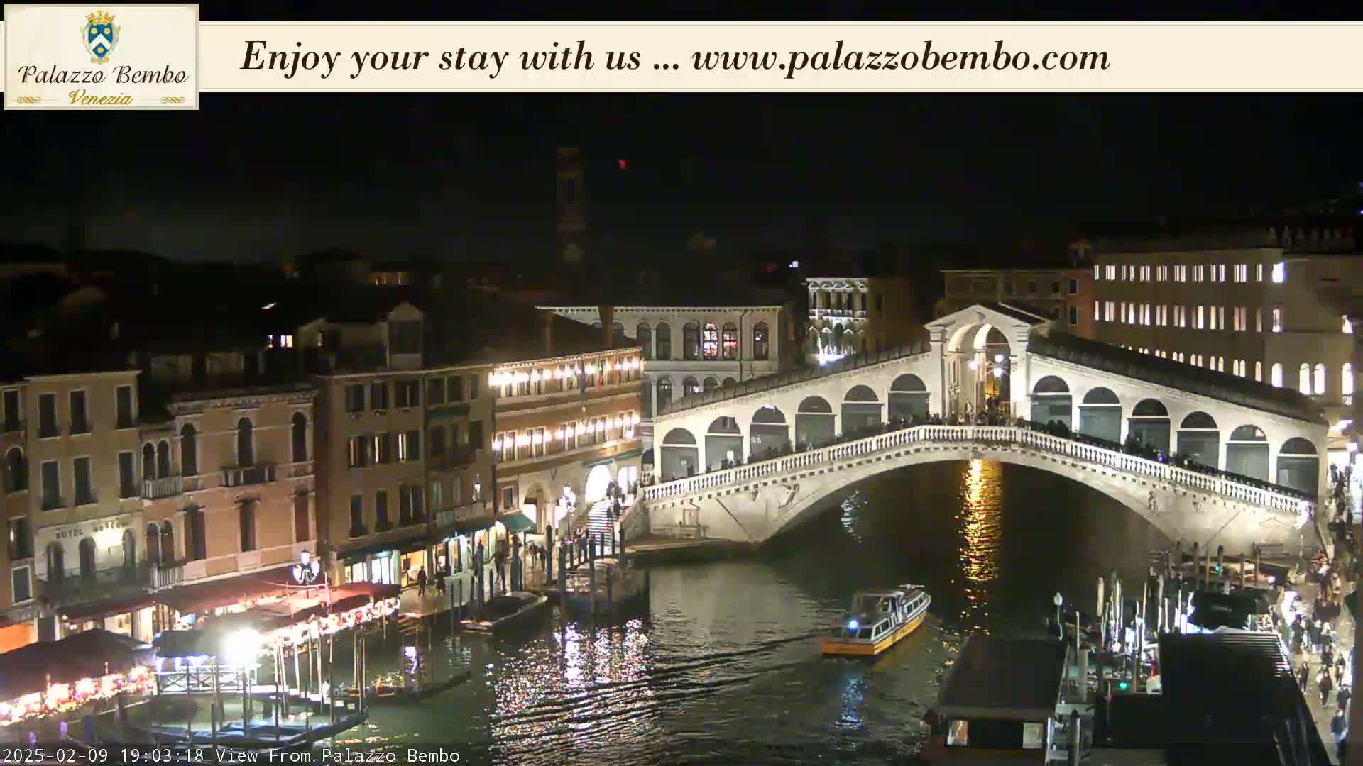 A nighttime view of a canal in Venice, showing a bridge with people walking across it and boats on the water.