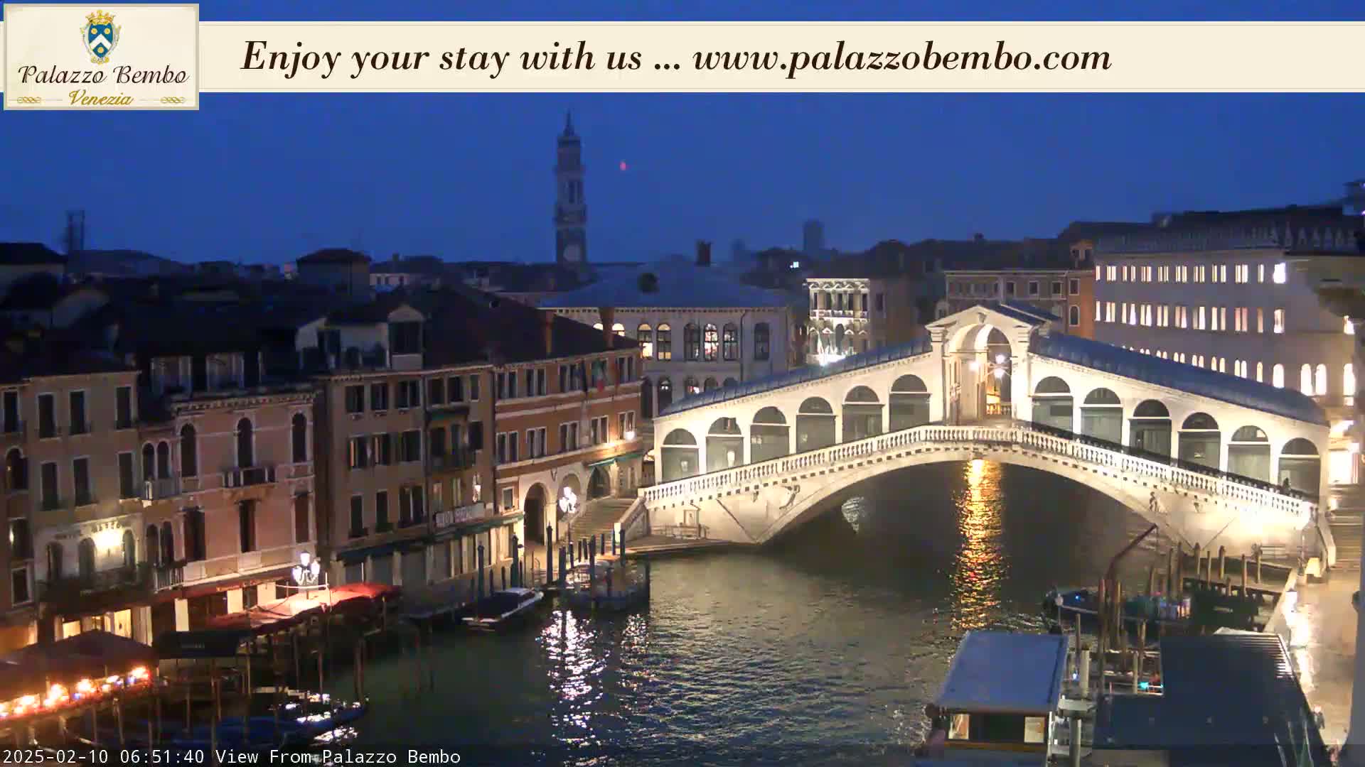 A nighttime view of a canal in Venice, showing a bridge lit up and buildings lining the waterway under a twilight sky.