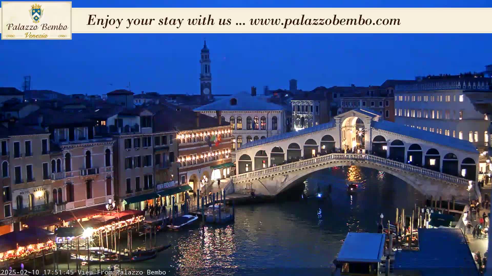 A nighttime view of a canal in Venice, with a bridge illuminated, buildings lining the waterway, and gondolas moored along the banks under a dark blue sky.