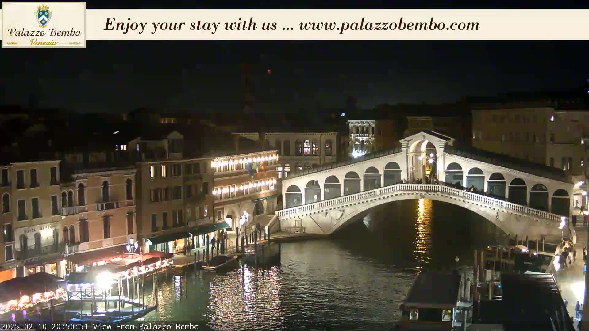 A nighttime view of a lit-up bridge spanning a canal in Venice, with buildings lining the waterway.