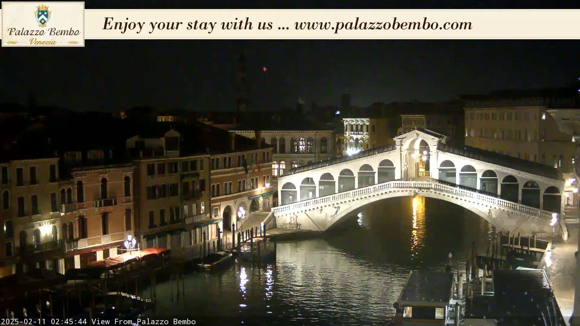 A nighttime view of a canal in Venice, showing a bridge lit up and buildings along the waterway under a clear, dark sky.
