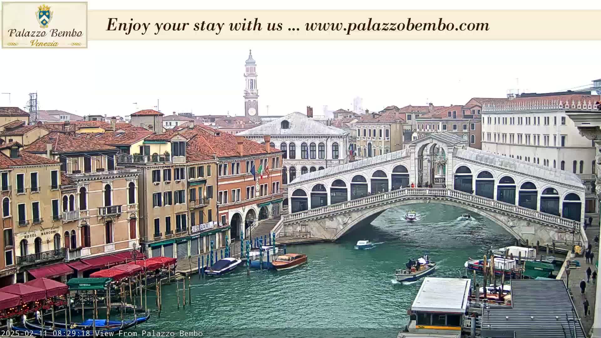 A view of a canal in Venice, Italy, shows a stone bridge with boats and buildings along its banks under an overcast sky.