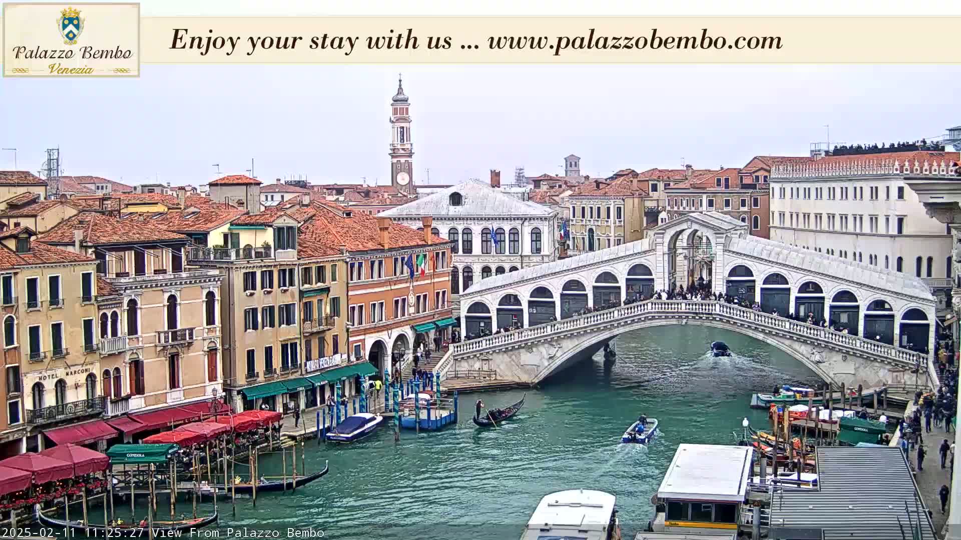 A canal in Venice, under an overcast sky, features the Rialto Bridge crowded with people, gondolas, and other boats, with buildings lining the waterway.