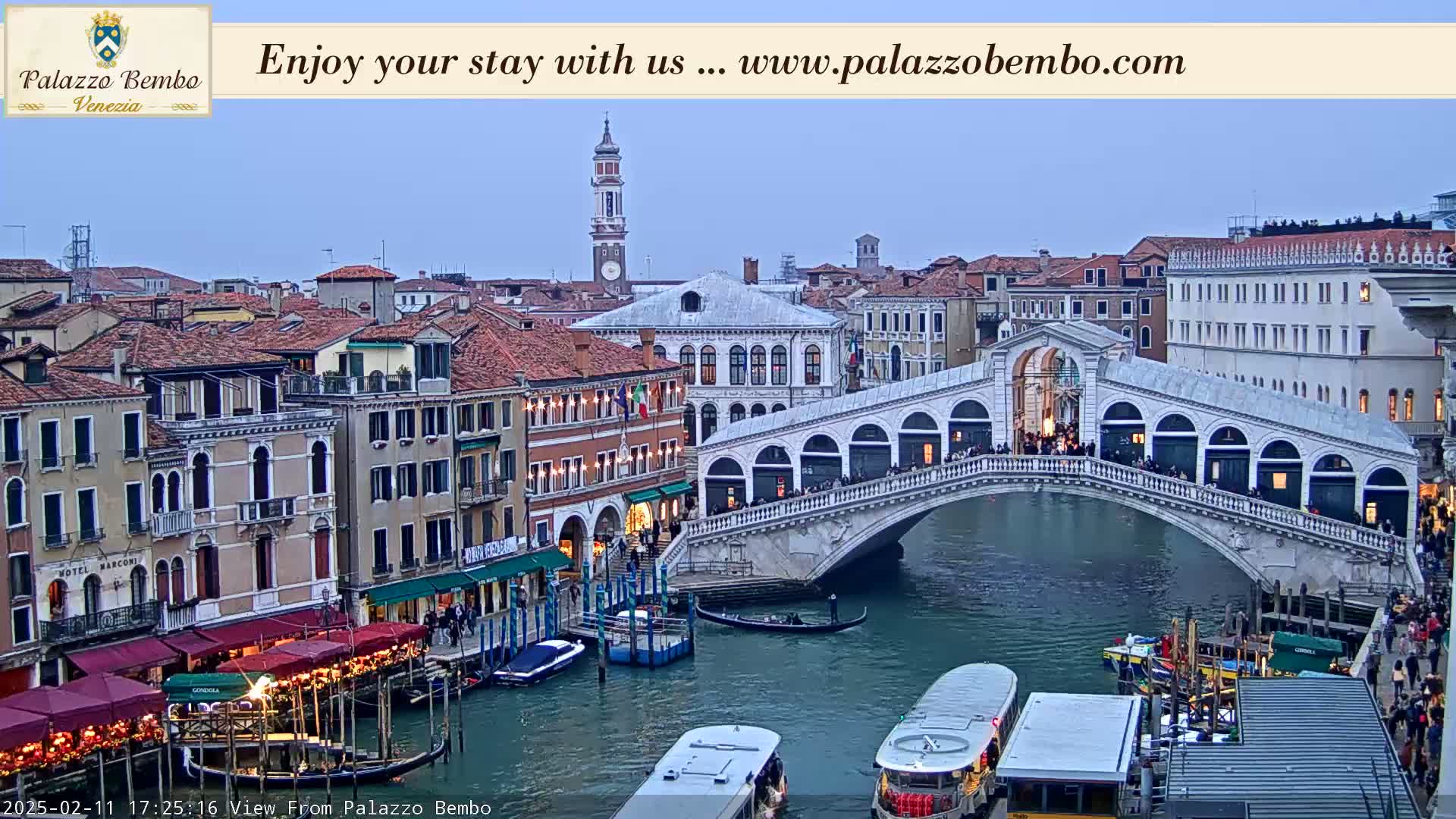 A view of a Venetian canal at twilight, showing a bridge with pedestrians and boats on the water, surrounded by buildings.