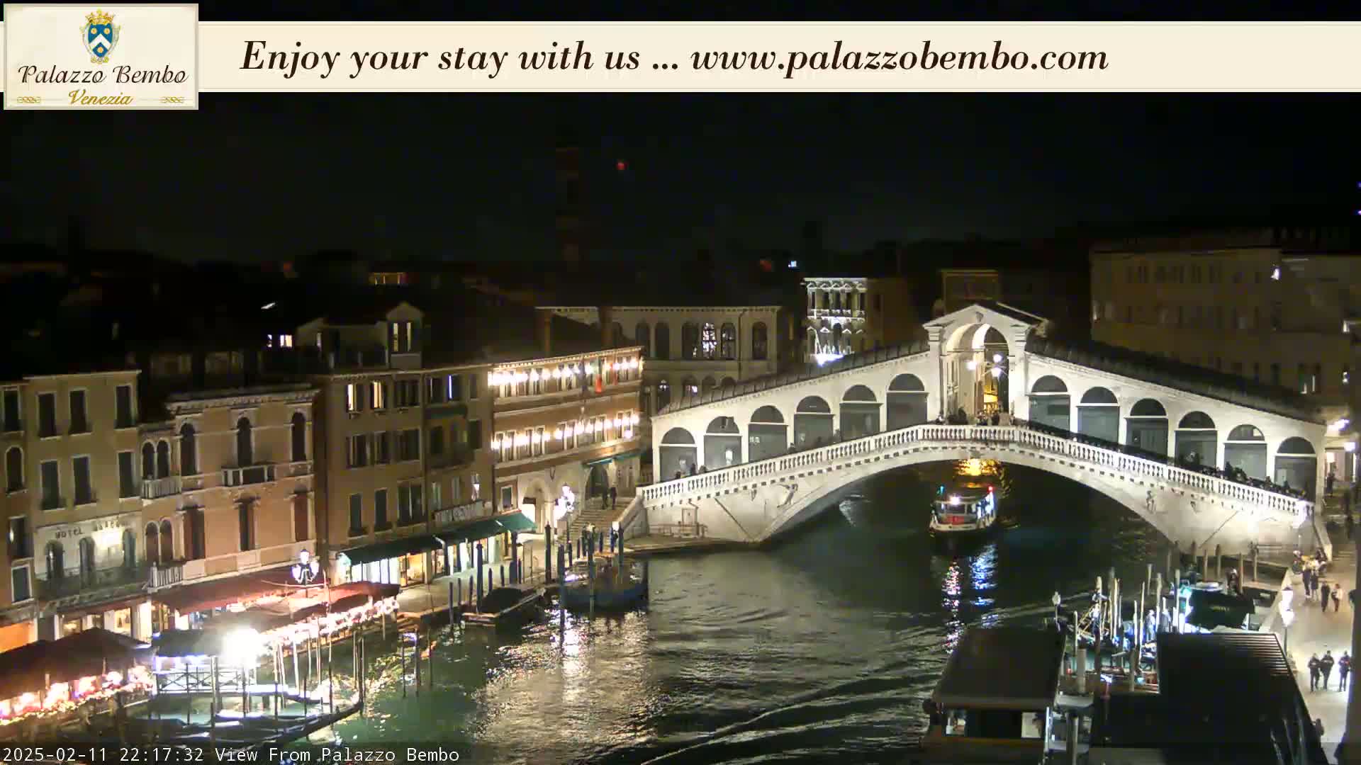 A nighttime view of a canal in Venice, showing a bridge illuminated and boats on the water under a clear, dark sky.