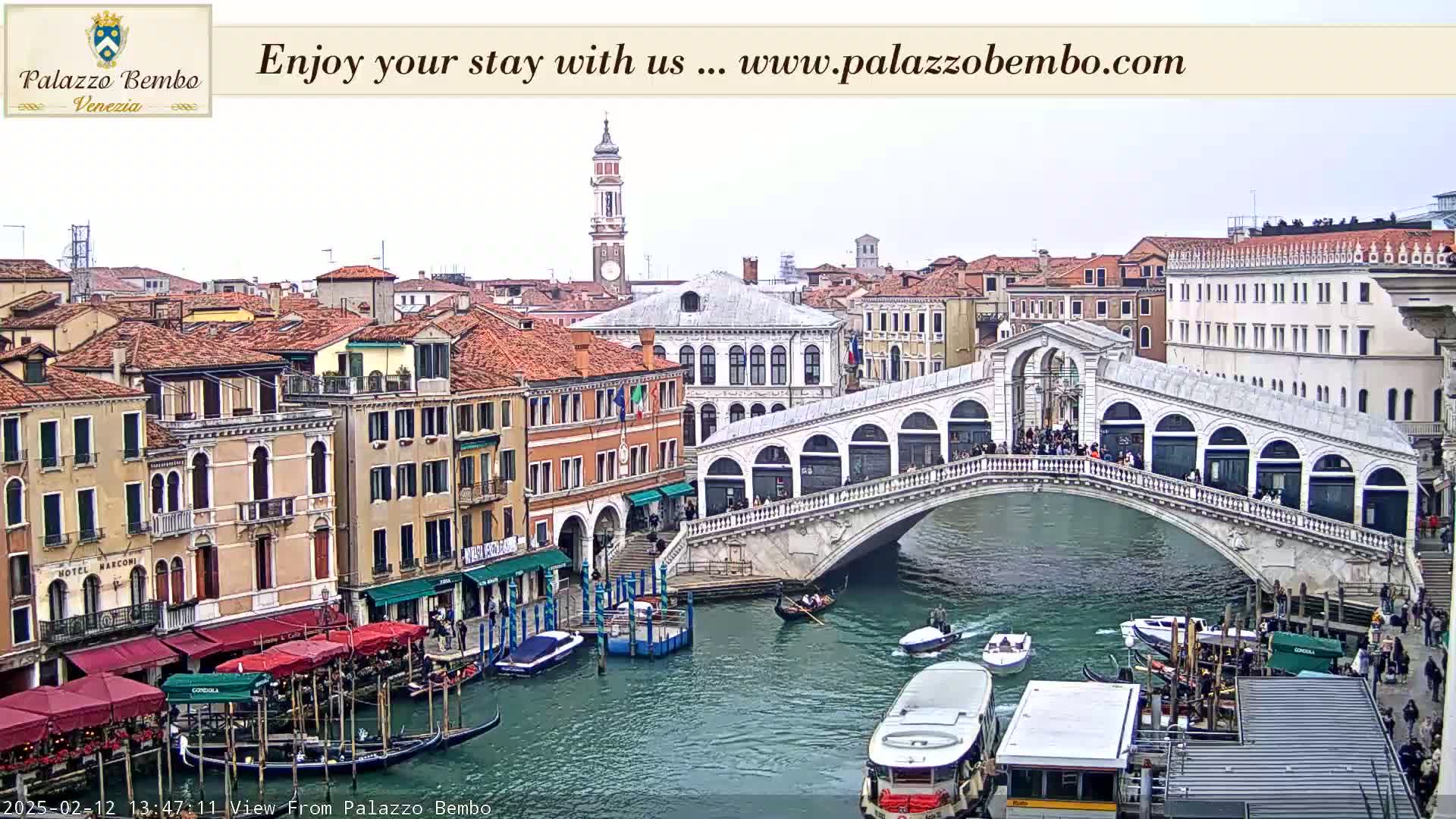 A view of a canal in Venice, under an overcast sky, shows a bridge with people walking across it and gondolas and other boats on the water, with buildings lining the canal.
