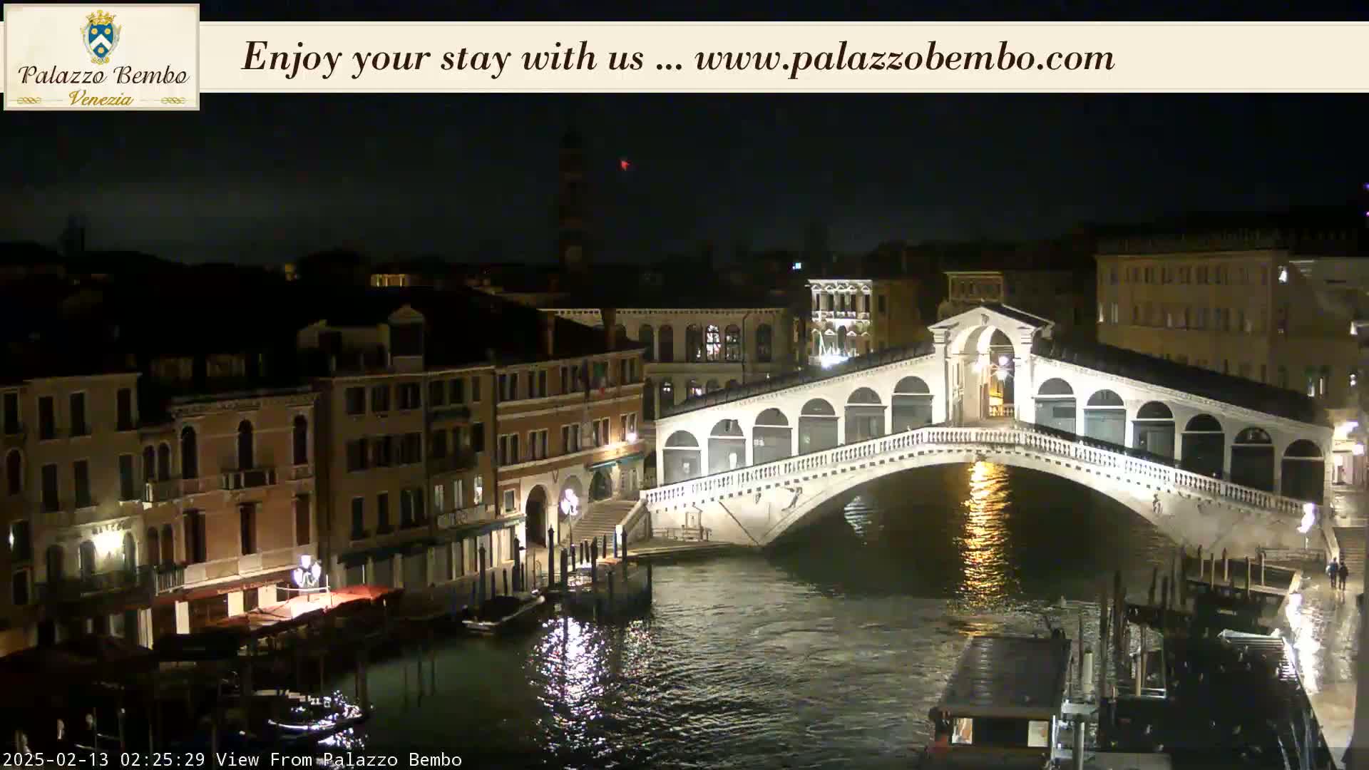 A nighttime view of a canal in Venice, Italy, shows the Rialto Bridge illuminated, with buildings lining the waterway.
