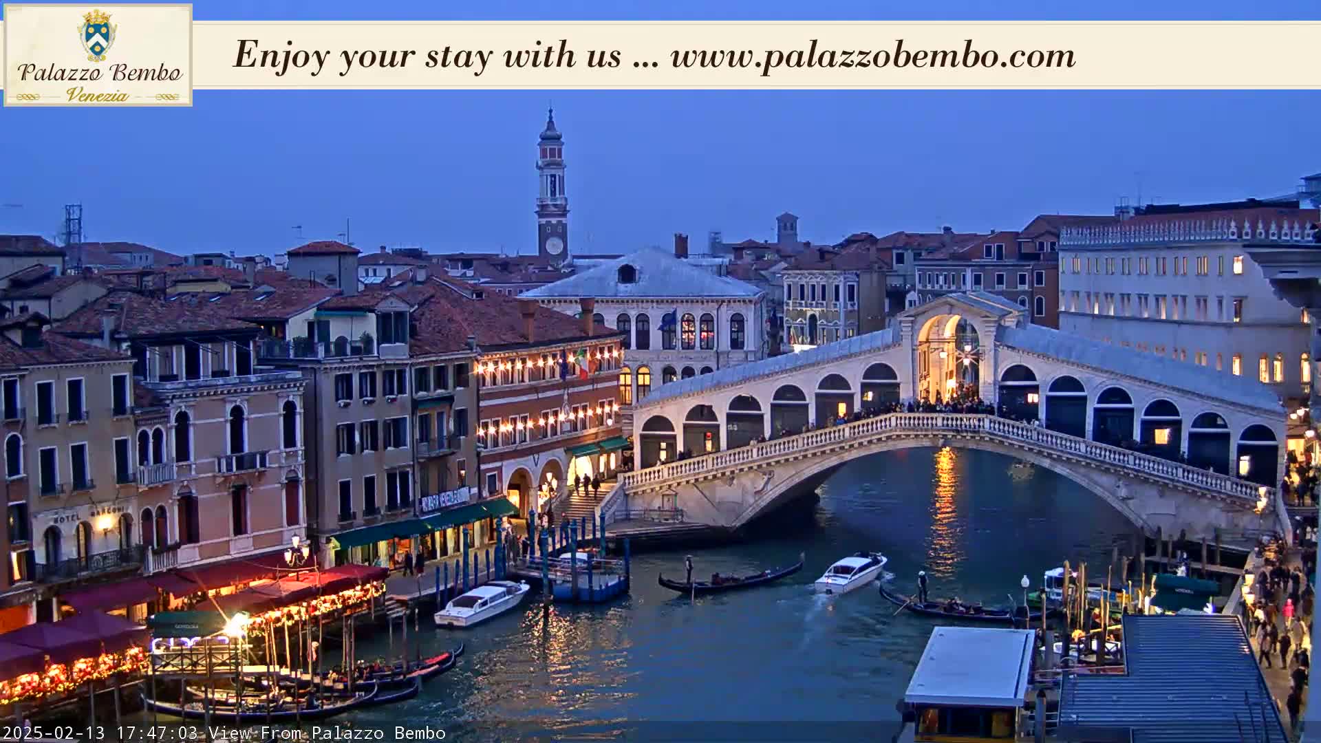 A nighttime view of a canal in Venice, showing the Rialto Bridge with gondolas and other boats on the water, under a twilight sky.