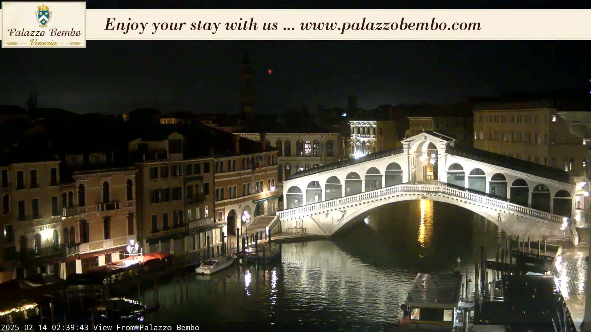 A nighttime view of a canal in Venice, showing a lit bridge and buildings along the water's edge under a dark sky.