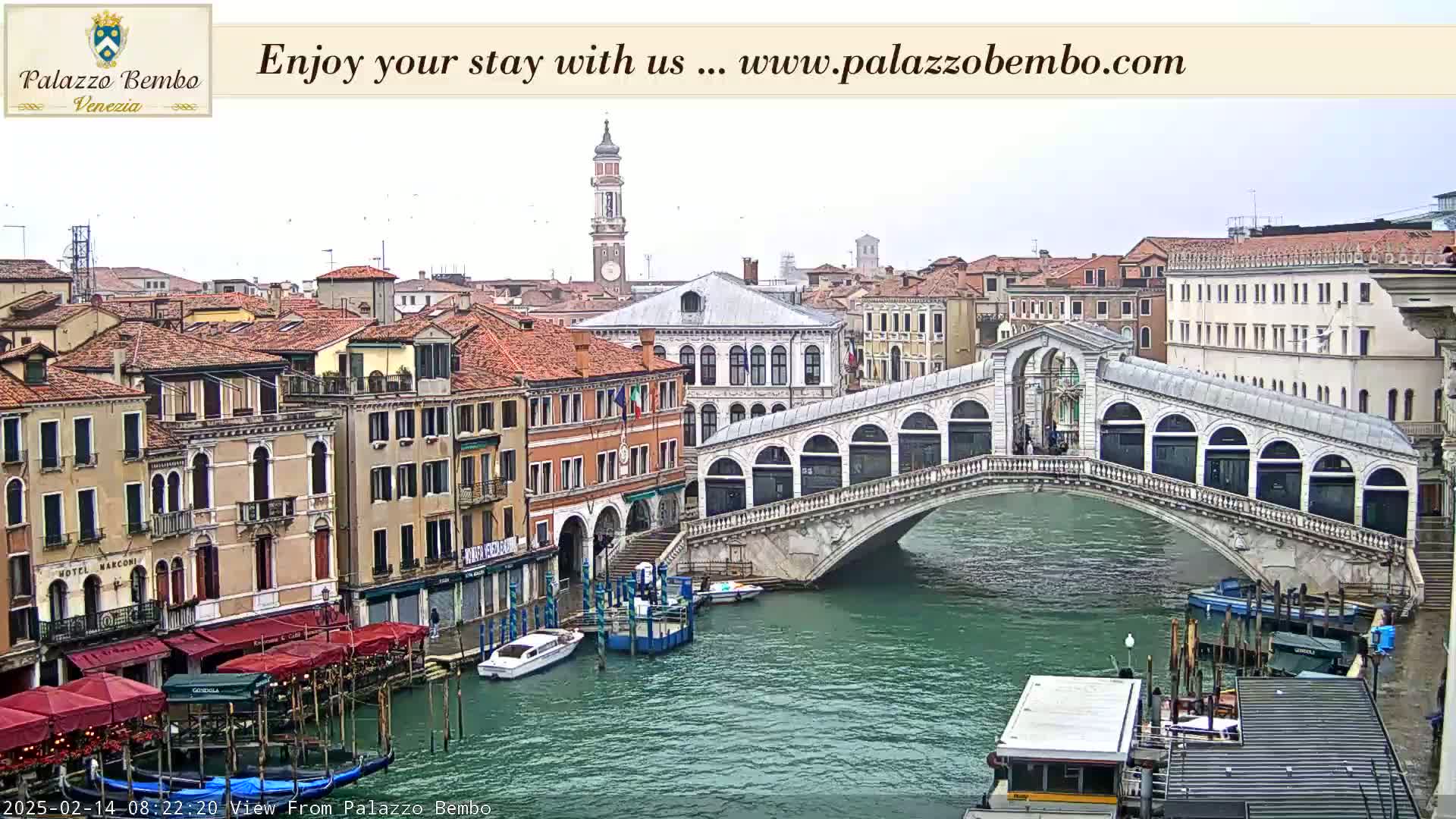 A view of a canal in Venice, overcast, showing a stone bridge, colorful buildings, and boats.