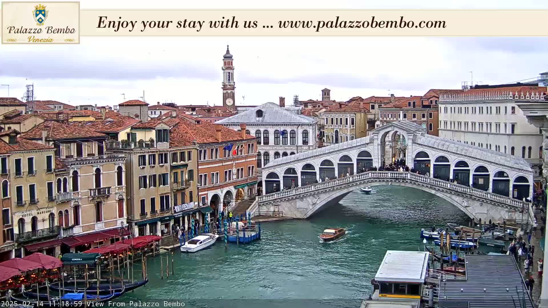 A view of a canal in Venice on an overcast day, showing a bridge with people walking across it and boats moored alongside buildings.