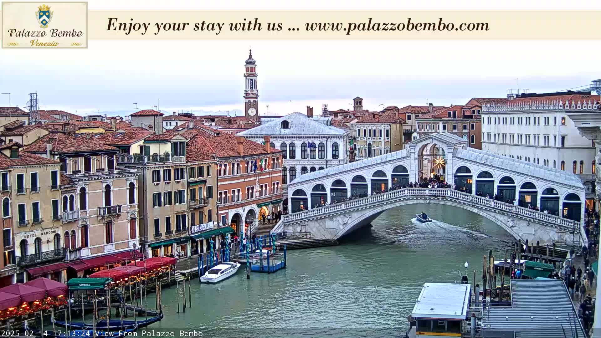A view of a canal in Venice, Italy, showing a bridge with people on it, boats on the water, and several buildings lining the canal under an overcast sky.