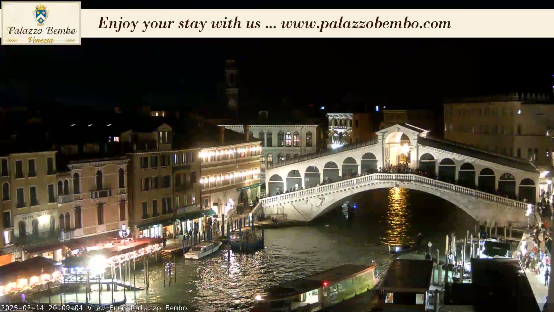 A nighttime view of a bridge over a canal in Venice, with illuminated buildings and boats lining the waterway.