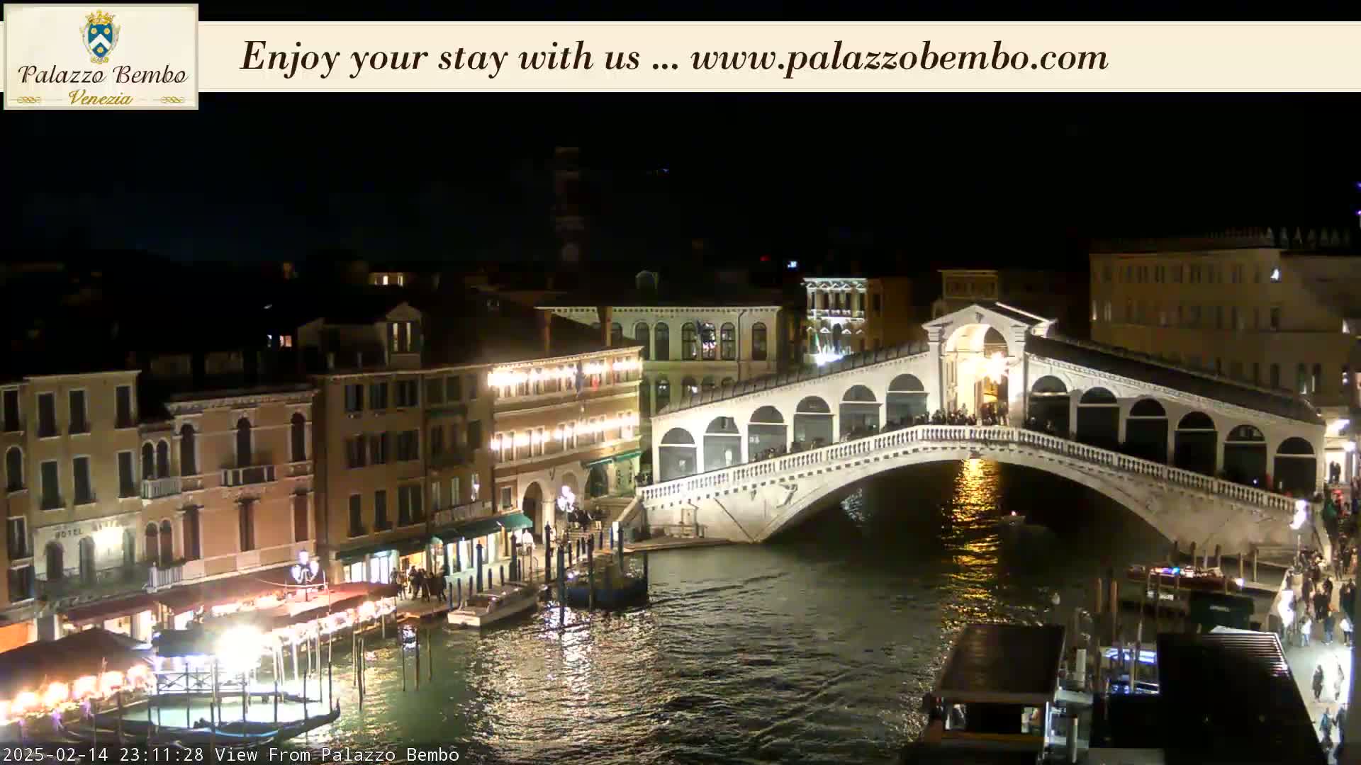 A nighttime view of a bridge over a canal in Venice, with illuminated buildings and boats along the waterway.