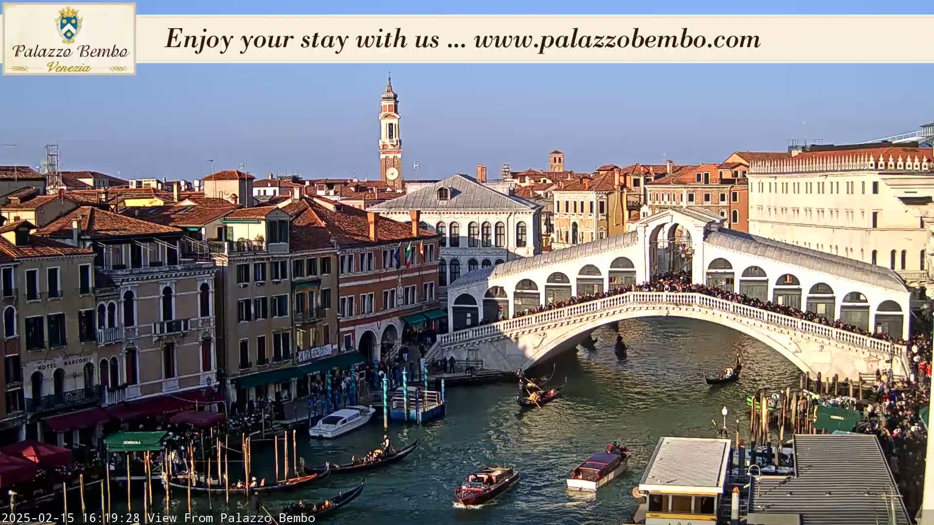 A sunny day in Venice shows a canal with gondolas and boats under the Rialto Bridge, which is crowded with people, with buildings lining the canal.