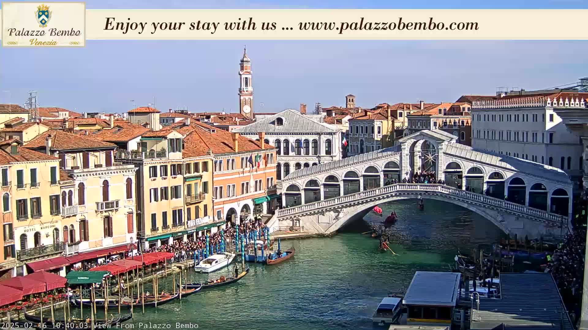 A sunny day in Venice shows a canal with gondolas, a bridge crowded with people, and numerous buildings along the waterfront.