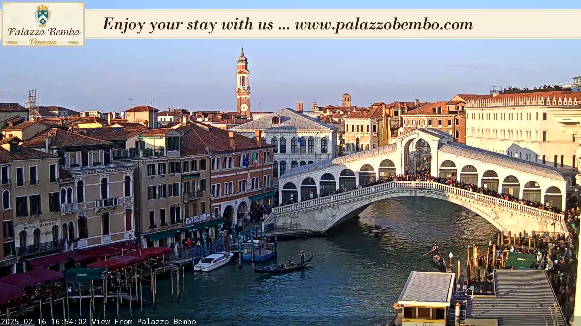 A Venetian bridge, crowded with people, arches over a canal lined with buildings under a clear, bright sky.