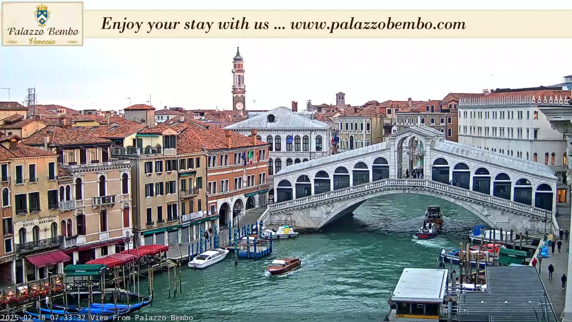 A view of a canal in Venice, under a mostly cloudy sky, shows a bridge with pedestrians crossing it and boats on the water, lined with buildings along the canal.