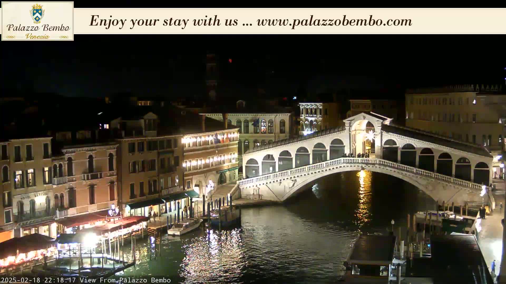 A nighttime view of a canal in Venice, showing a bridge illuminated, with buildings lining the waterway.