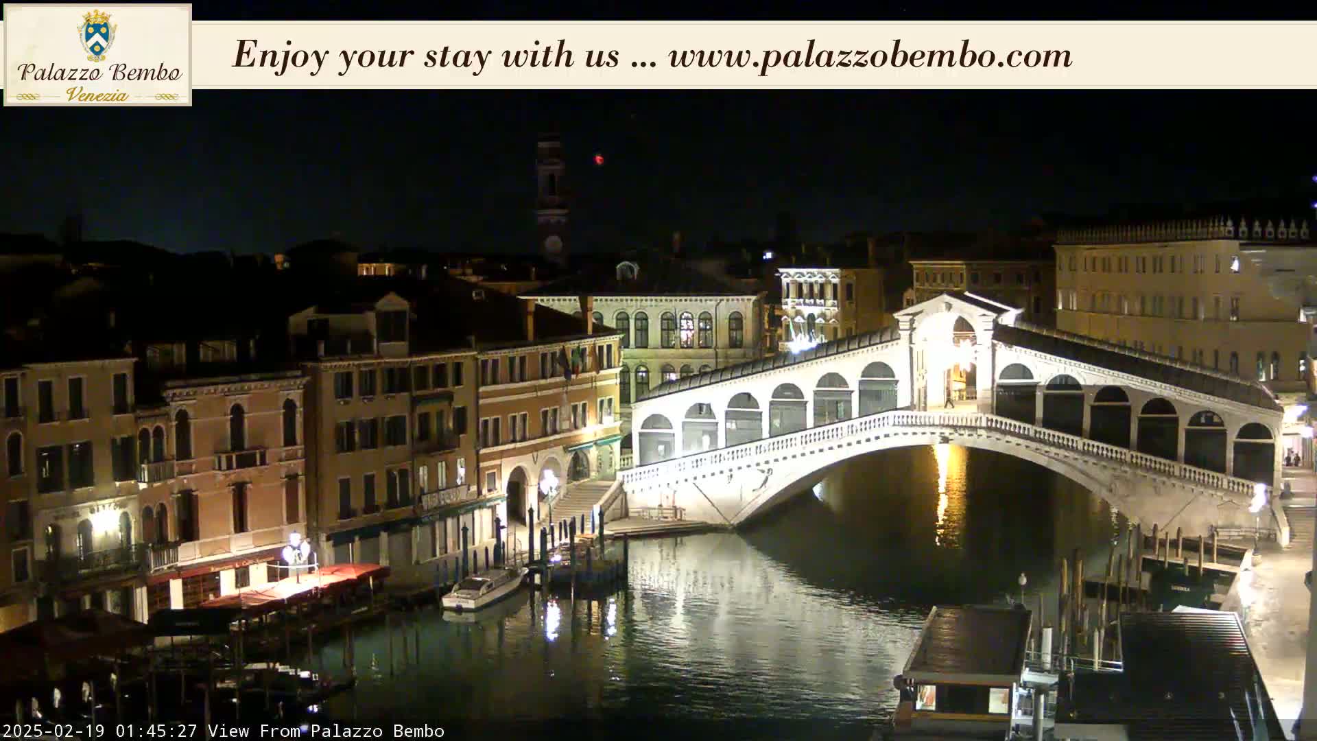 A nighttime view of a bridge over a canal in Venice, with buildings lit along the waterway.