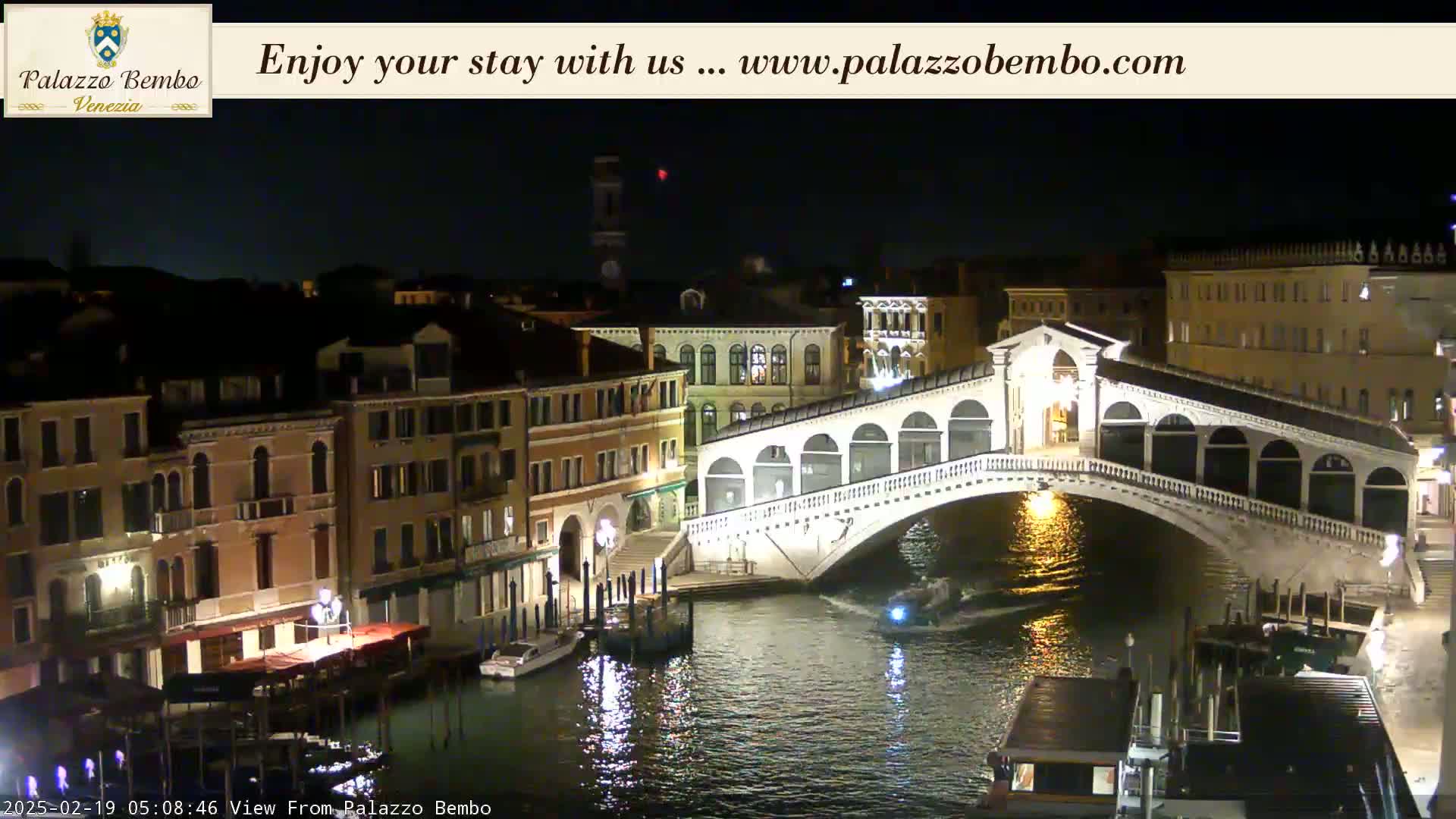A nighttime view of a canal in Venice, showing the Rialto Bridge lit up, with boats on the water and buildings lining the canal.