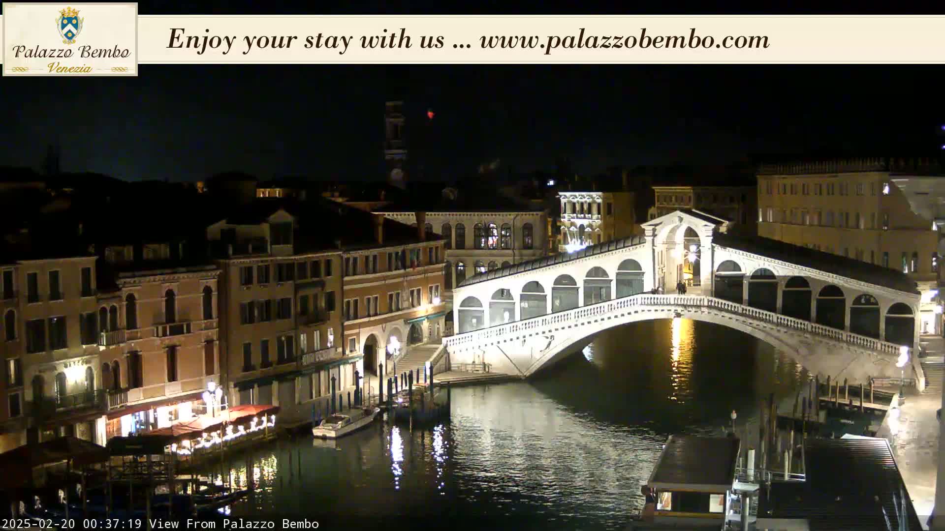 A nighttime view of a canal in Venice, showing a bridge lit up and buildings lining the waterway under a clear, dark sky.