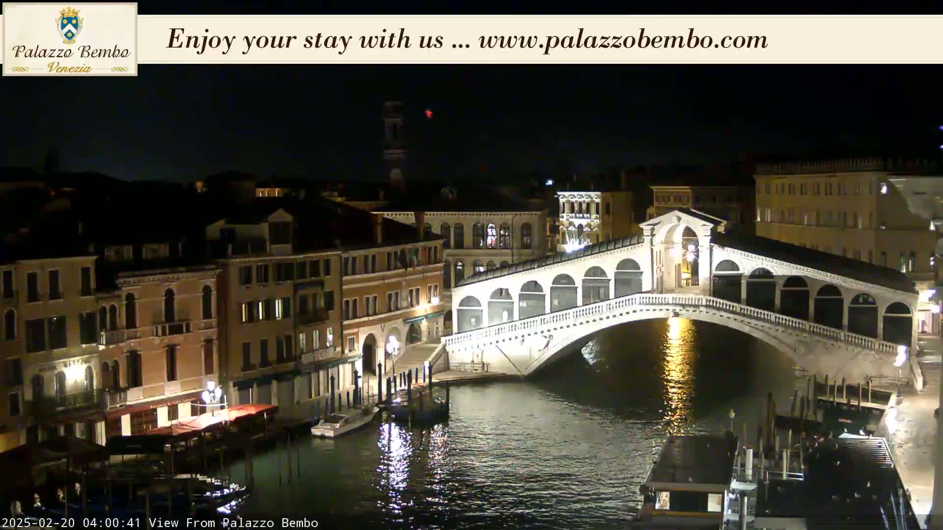 A nighttime view of a canal in Venice, with the Rialto Bridge illuminated and boats moored along the waterway.