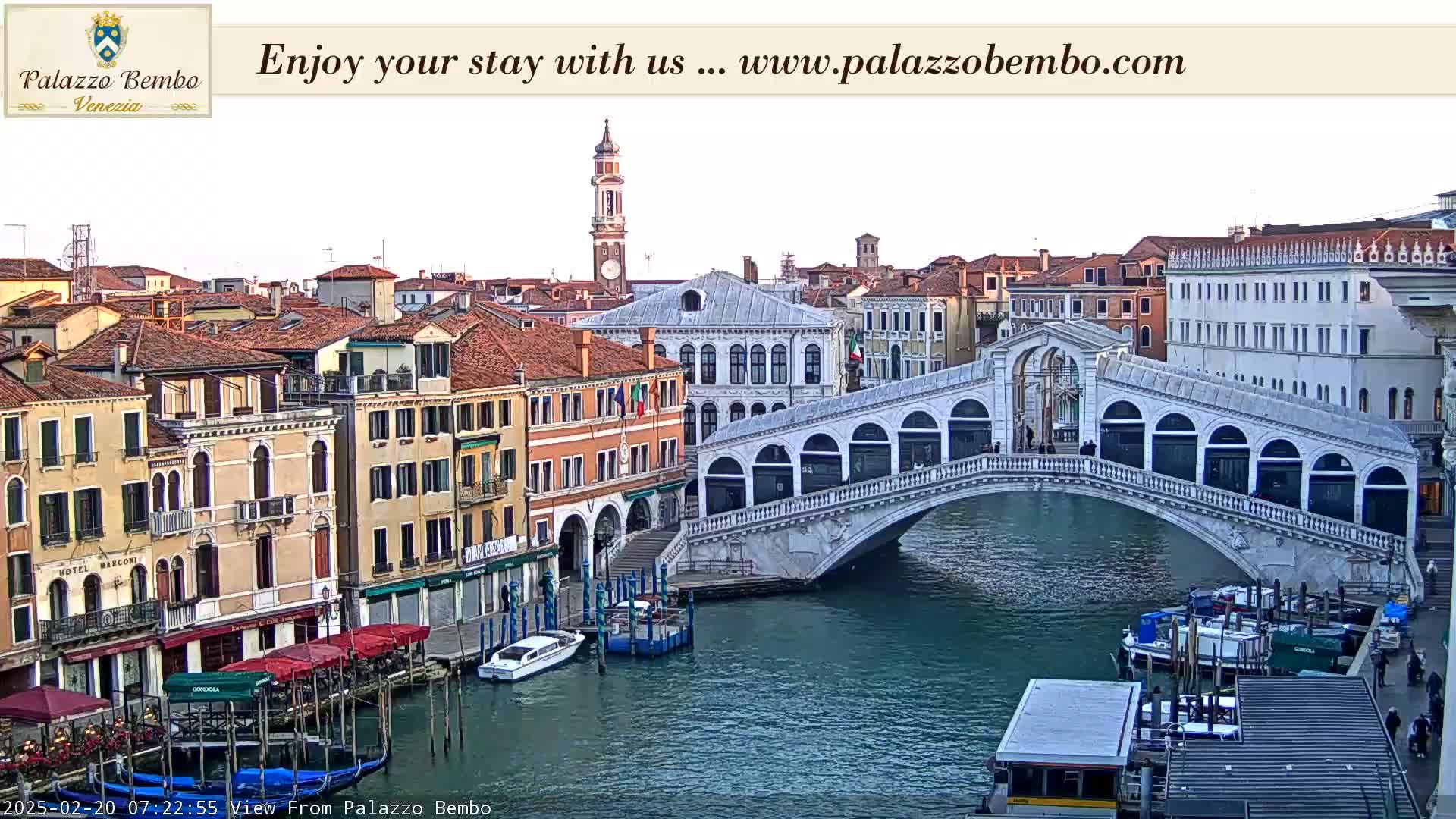 A view of a canal in Venice, Italy, shows a bridge with boats moored alongside and buildings lining the waterway under a mostly clear sky.