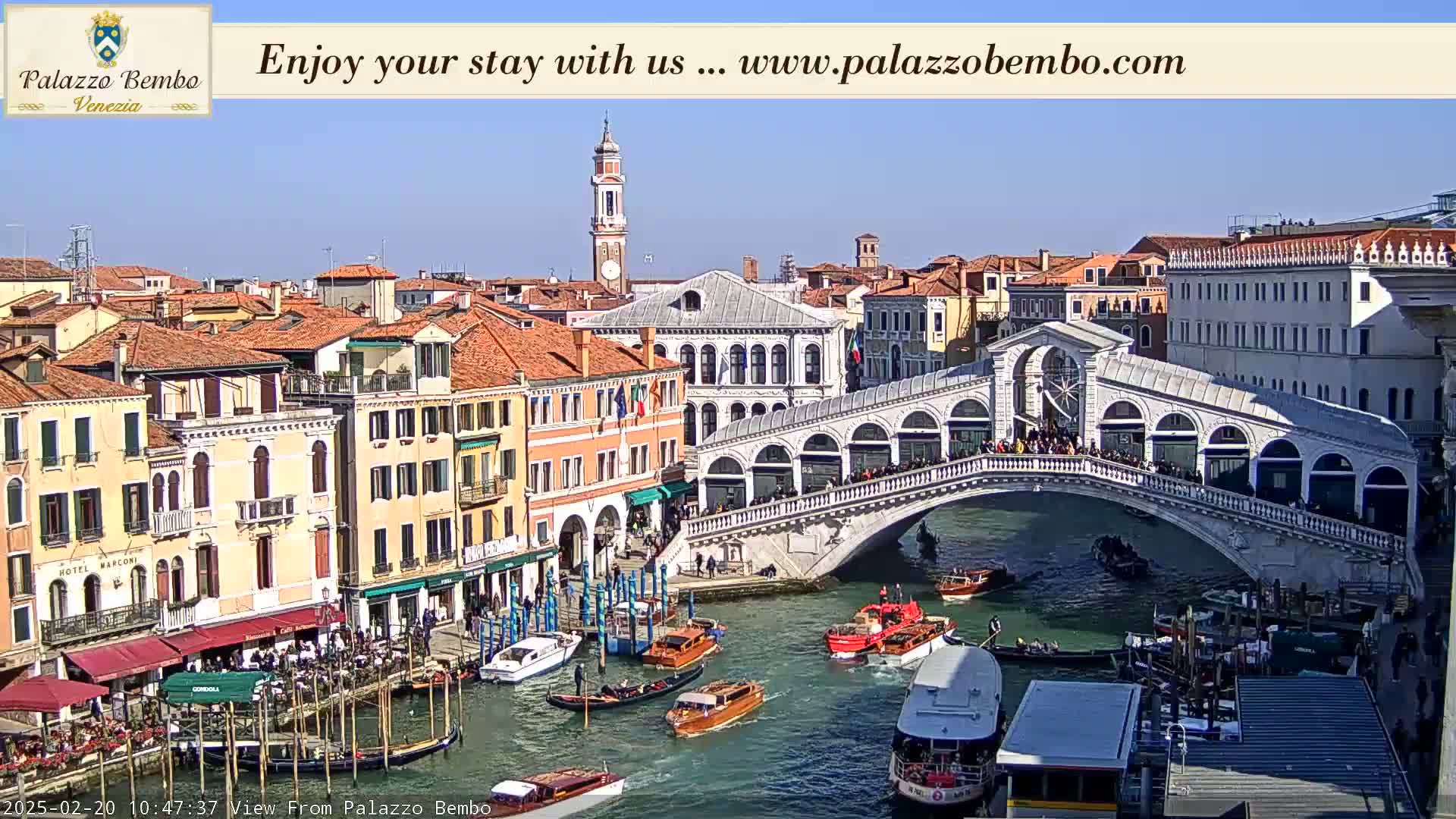 A sunny view of a canal in Venice, Italy, shows the Rialto Bridge crowded with people, boats on the water, and many buildings lining the waterway.