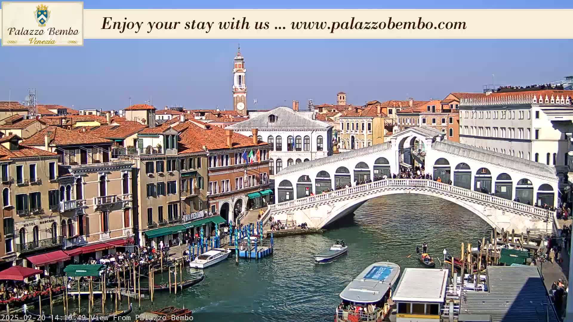 A sunny view of a canal in Venice, with a bridge crowded with people, boats, and buildings lining the waterway.