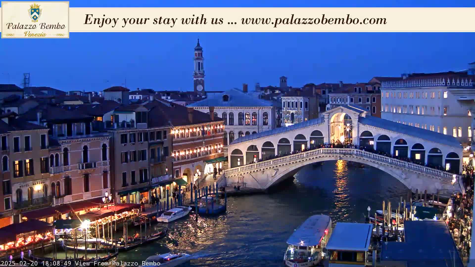 A twilight view of a canal in Venice, showing a bridge with people on it and buildings lit up along the waterway.
