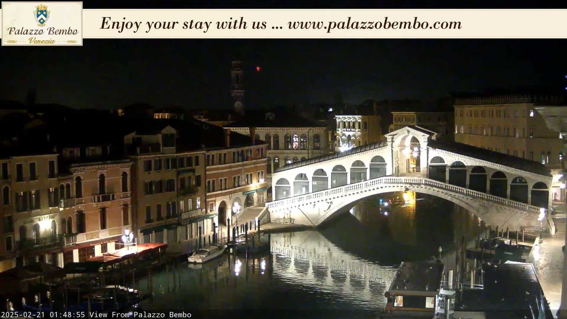 A nighttime view of a canal in Venice, showing a bridge and buildings lit up along the waterway under a clear, dark sky.