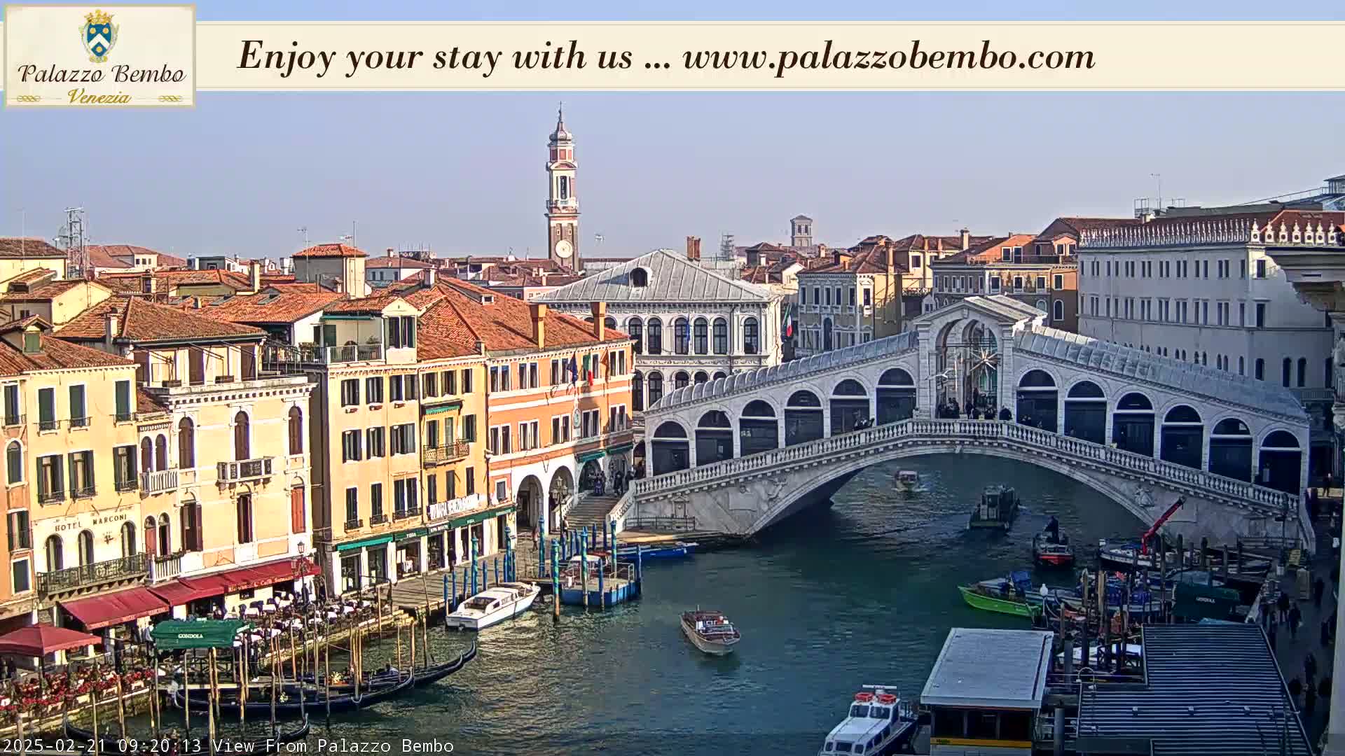 A sunny view of the Rialto Bridge in Venice, Italy, with boats on the canal and buildings lining the waterways.