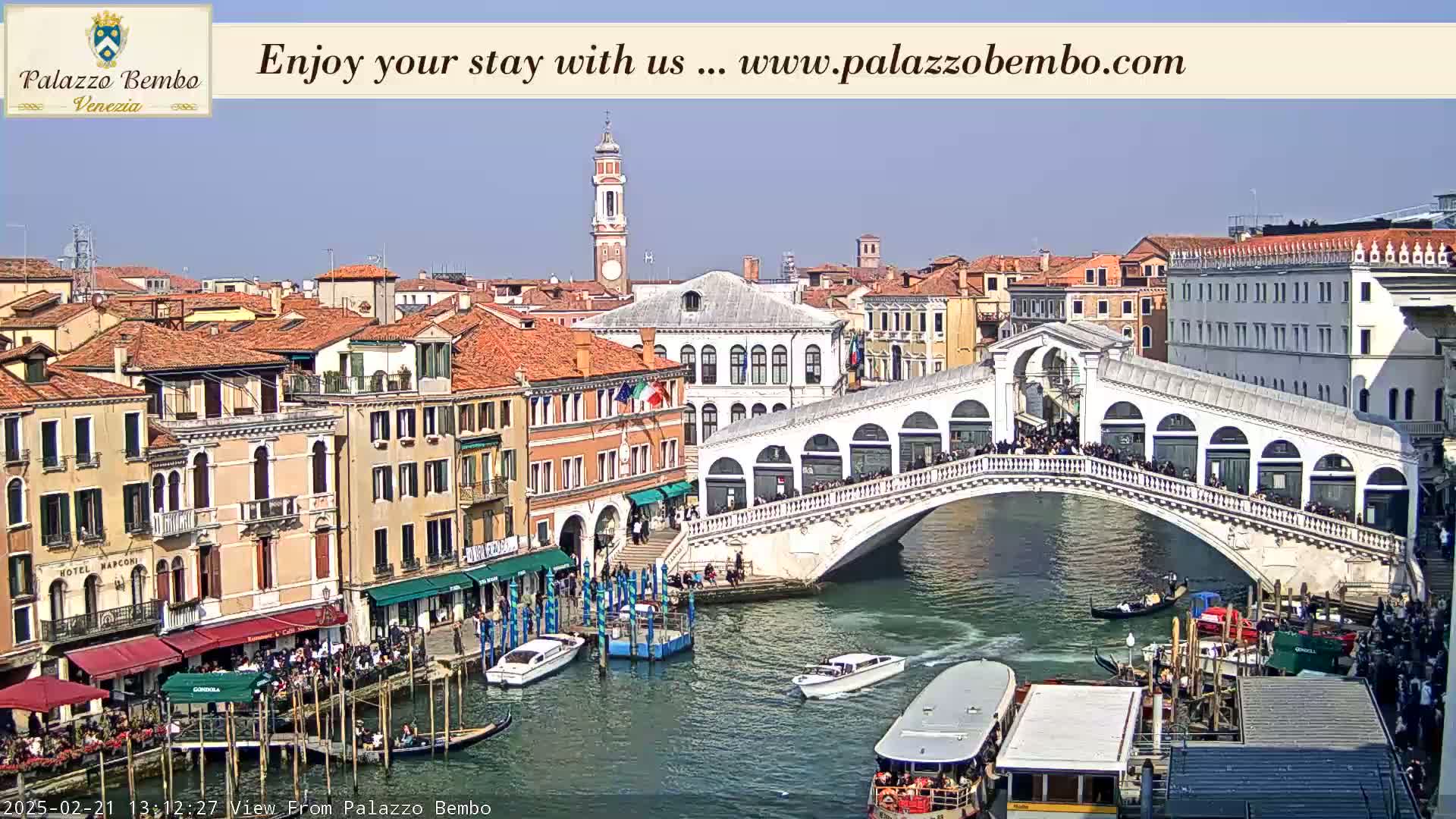 A sunny day in Venice shows a canal with boats, a bridge crowded with people, and buildings lining the waterway.
