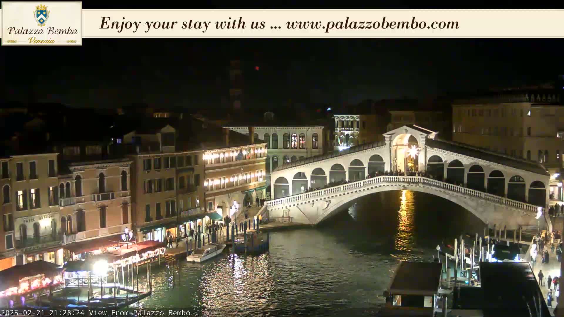 A nighttime view of a canal in Venice, showing a bridge lit up with people walking across it and buildings lining the waterway.