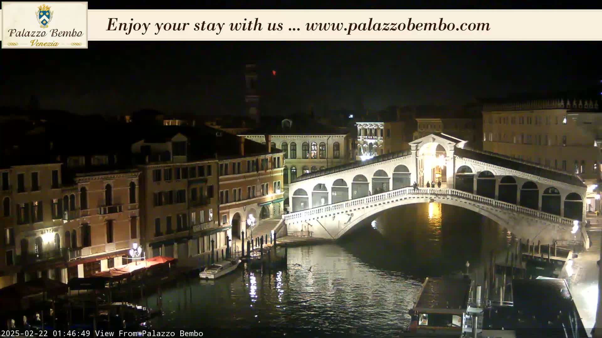 A nighttime view of a canal in Venice, showing a bridge lit up with lights reflecting on the water.
