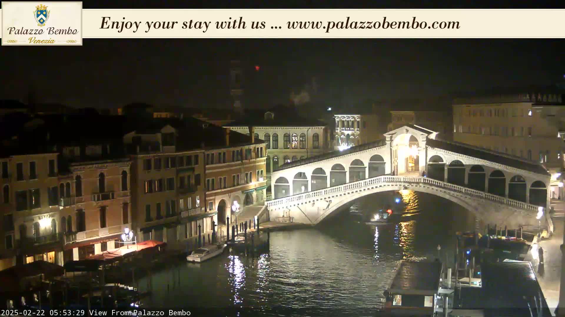 A nighttime view of a canal in Venice, showing a bridge lit up and boats on the water under a dark sky.